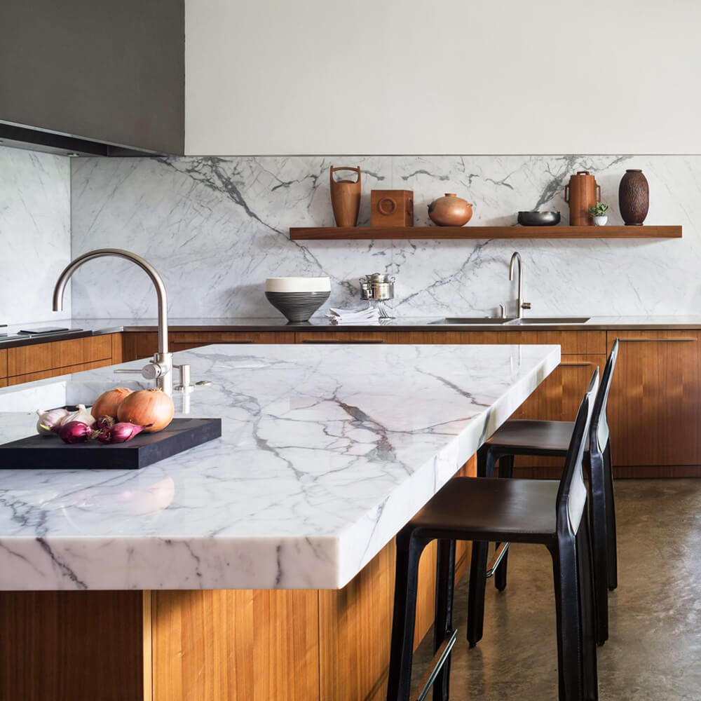 Modern kitchen featuring a striking marble waterfall island and backsplash, paired with warm wooden cabinetry, minimalist shelving, and sleek bar seating.