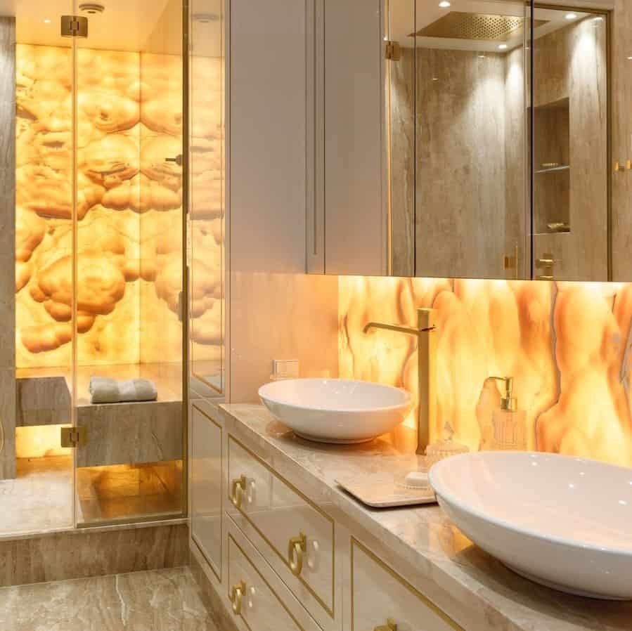 Luxurious bathroom interior with illuminated Honey Onyx marble backsplash and shower wall, featuring dual white vessel sinks on a beige vanity unit.