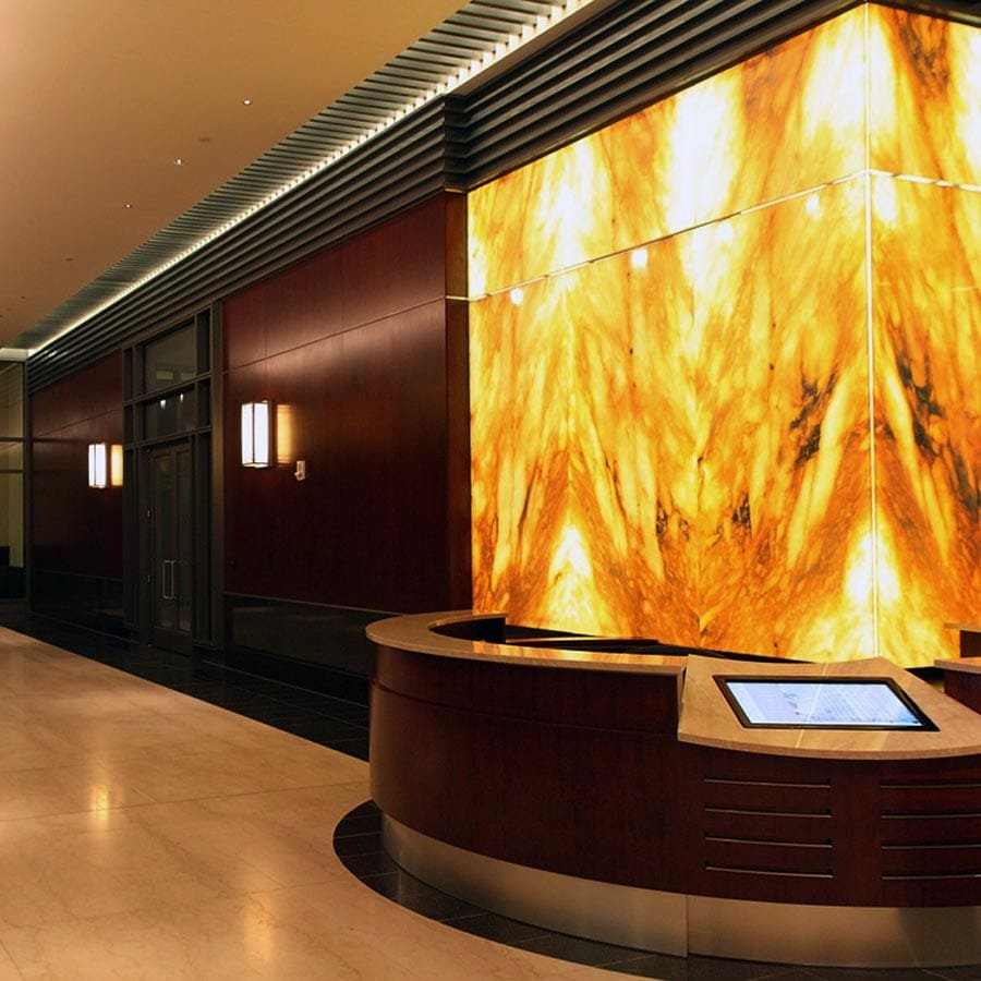 Elegant hotel reception area featuring a stunning illuminated Honey Onyx stone wall panel with bold amber and golden tones, complemented by dark wood finishes and modern ambient lighting.