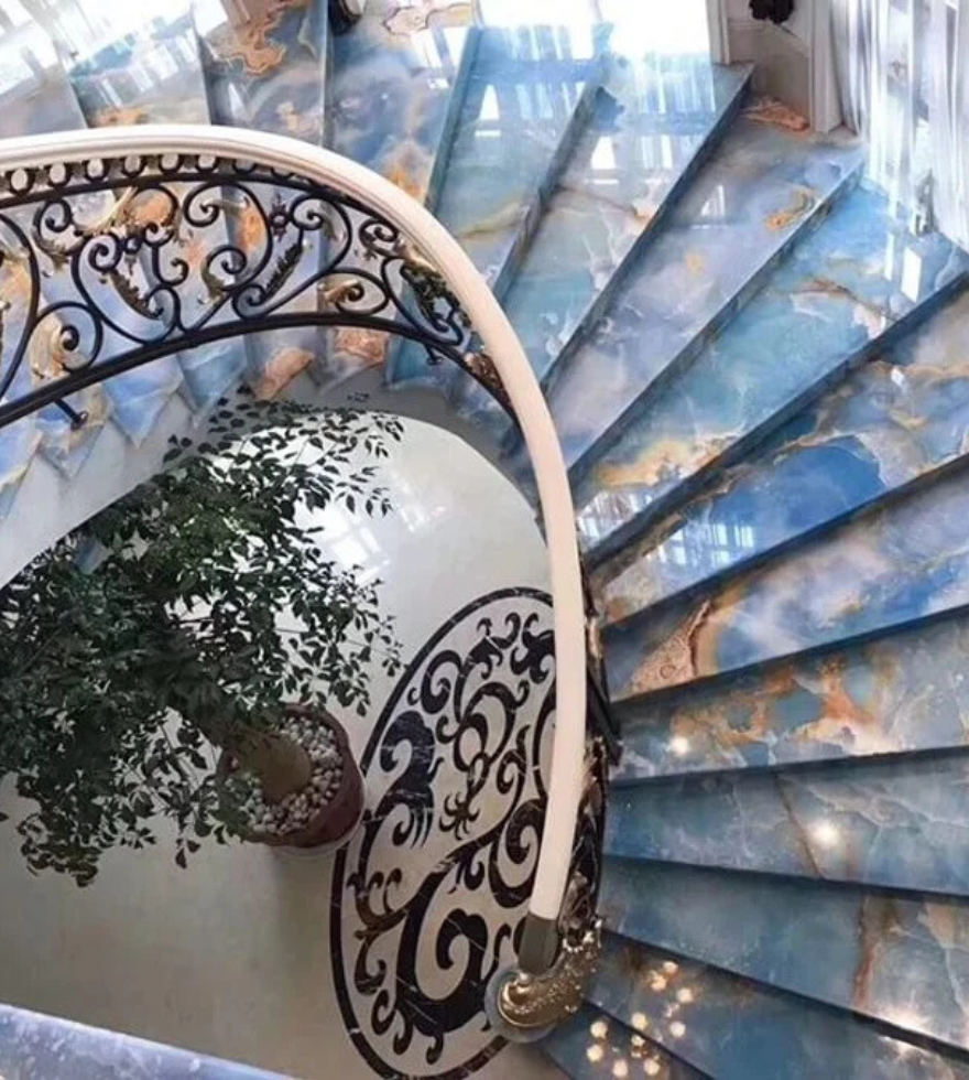 Elegant spiral staircase clad in Blue Onyx marble with golden veining, featuring ornate wrought iron railings and a central indoor plant.
