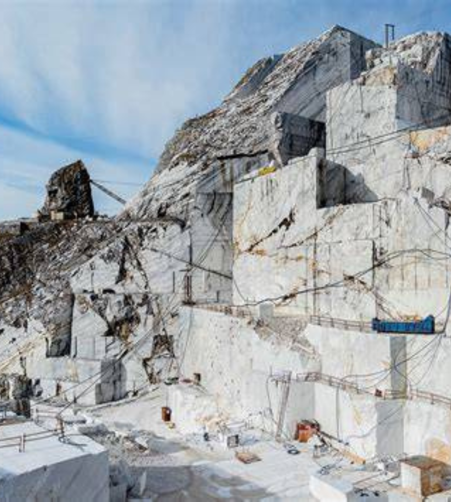 White marble quarry set against a mountainous backdrop, showcasing large cut stone blocks, rugged terrain, and industrial equipment under a clear blue sky.