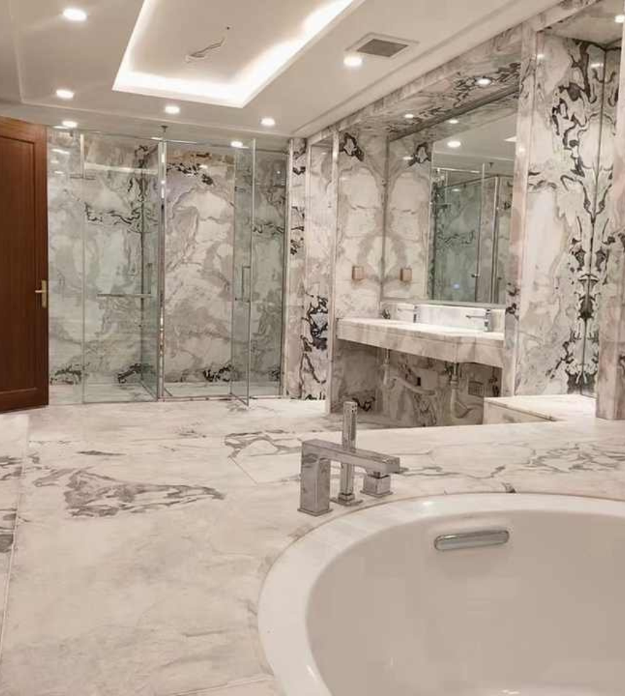 Opulent bathroom interior fully clad in luxurious White Dover Marble, featuring bold black veining, a soaking tub, expansive mirrors, and ambient recessed lighting.